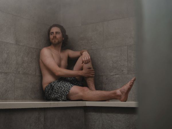 A shirtless man lounges in a tiled bathtub, propped up against the wall with his legs out and arms resting.