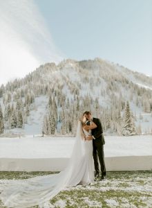 A bride and groom in wedding attire kiss outdoors by a snowy field, with evergreen trees and mountains in the background.