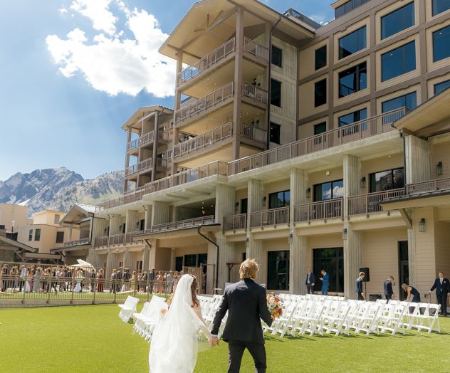 A couple walks toward a ceremony on a lawn beside a modern hotel, with wedding chairs and guests under sunny skies.