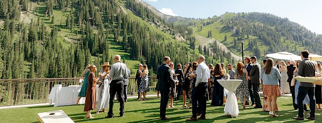 An outdoor wedding reception on a sunny lawn with guests mingling, overlooking green alpine mountains and towering clouds.