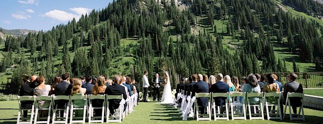 An outdoor wedding ceremony on a grassy lawn with guests seated and a couple facing a backdrop of mountains and trees.