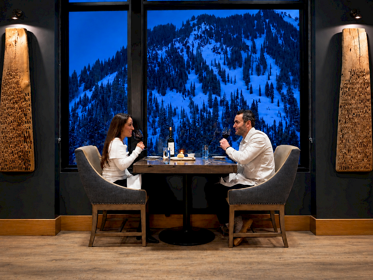 Couple toast wine in a cozy restaurant, with a snowy pine-forest view outside.