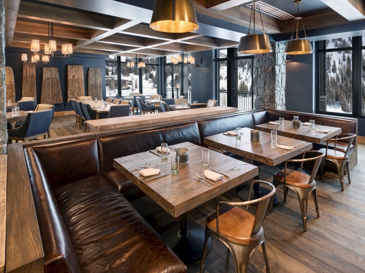 A cozy mountain restaurant dining area with wooden tables, leather booths, pendant lights, and snowy views through large windows.