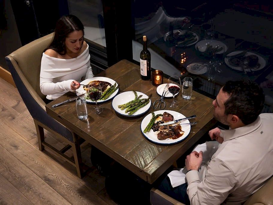 A man and woman dine by candlelight at a restaurant table with plated meals, asparagus, and a bottle of red wine.