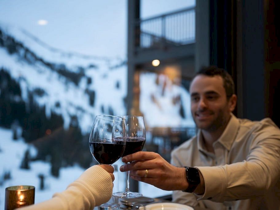 Two people clink red wine glasses in a cozy restaurant by a window overlooking snow-covered scenery.