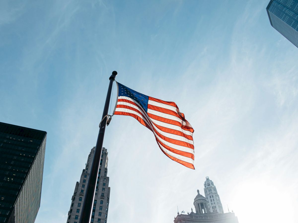 The American flag waves between tall skyscrapers under a blue sky, with city buildings framing the view.