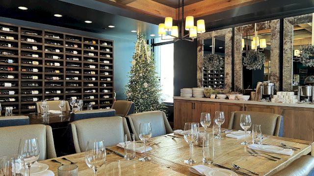A restaurant dining room with a tall decorated Christmas tree, a chandelier, and walls packed with wine bottles.