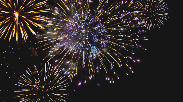 Colorful fireworks burst across a dark night sky, with gold, blue, and pink trails lighting up the scene.