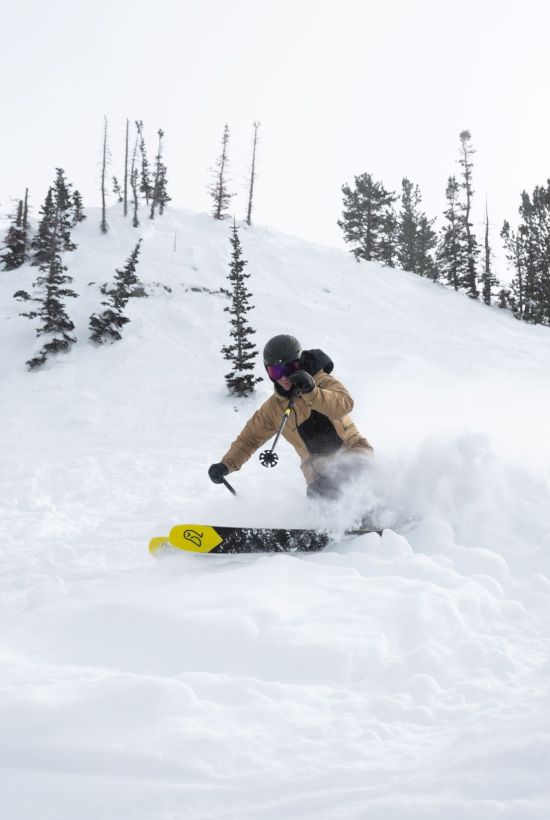 A snowboarder in goggles carves down a snowy mountain, kicking up a cloud of powder as pine trees line the slope.