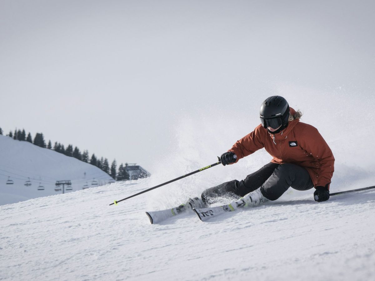 A skier in a helmet carves down a snowy mountain slope near ski lifts, kicking up powder as they speed downhill.