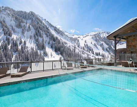 A heated outdoor pool beside a wooden deck with loungers, overlooking snowy alpine mountains under a clear blue sky.