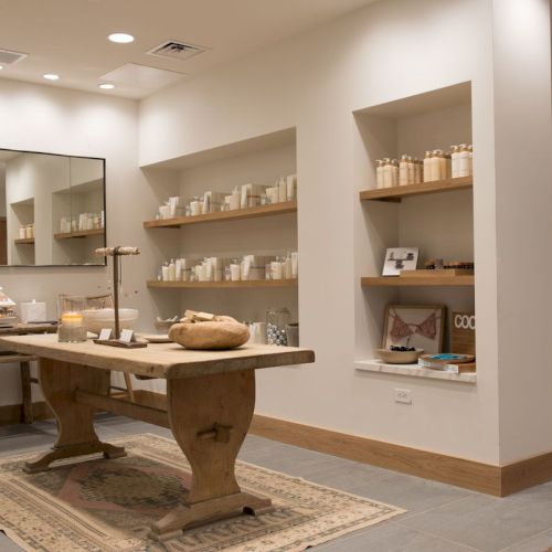 A bright spa/retail room with candles, shelves of skincare bottles, and a wooden treatment table centered on rugs.