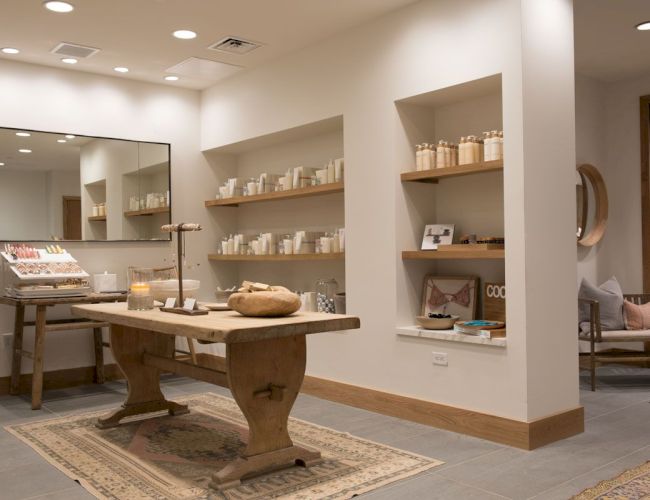 A bright spa/retail room with candles, shelves of skincare bottles, and a wooden treatment table centered on rugs.