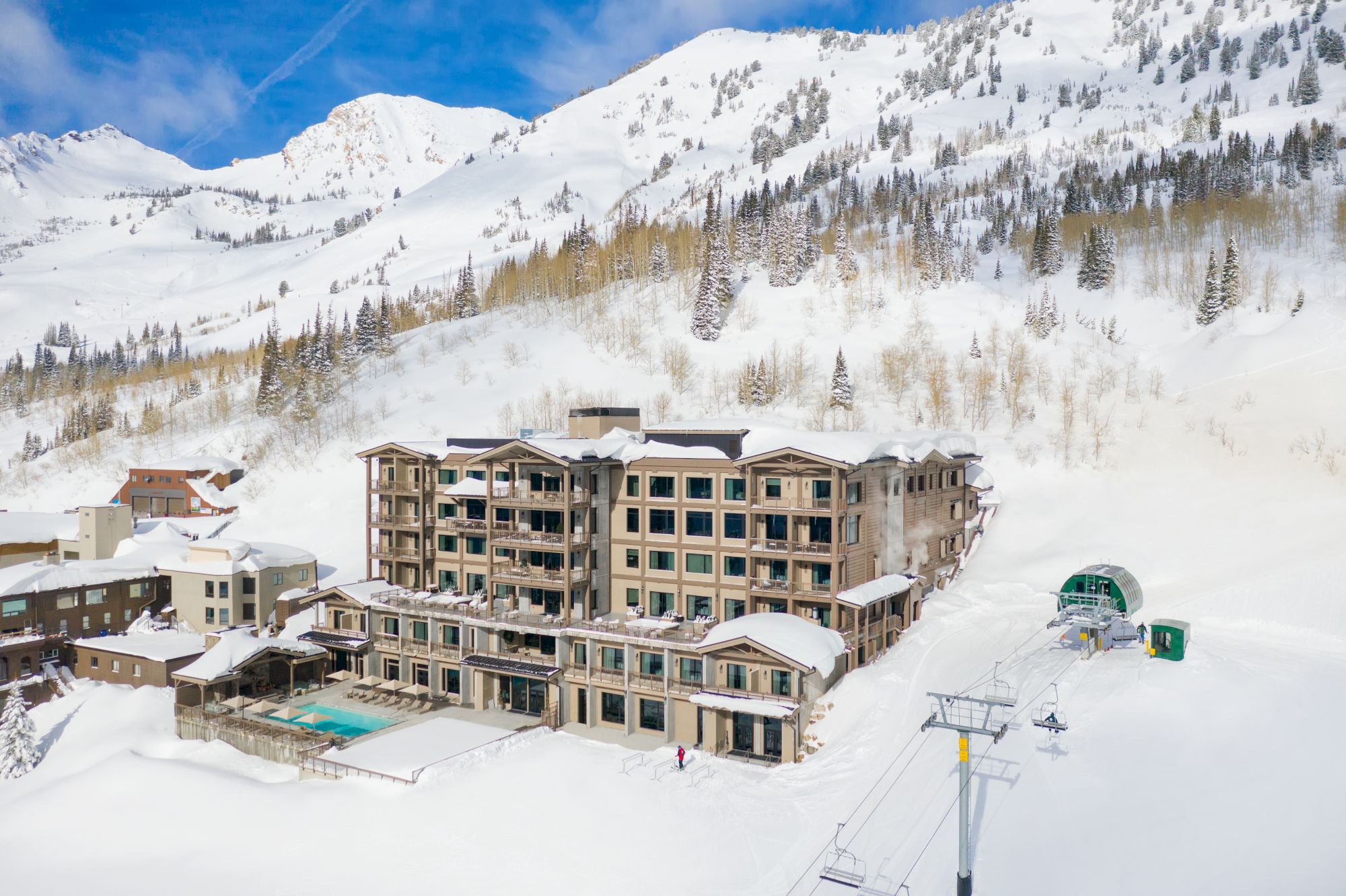 Snowy mountain resort with a large hotel, pine trees, and ski lifts below towering Alps under a bright blue sky.