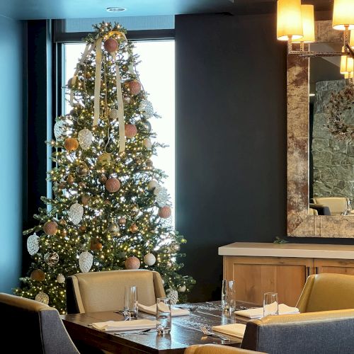 A decorated Christmas tree with warm lights stands in a restaurant dining area, beside wine bottles and a stone fireplace.