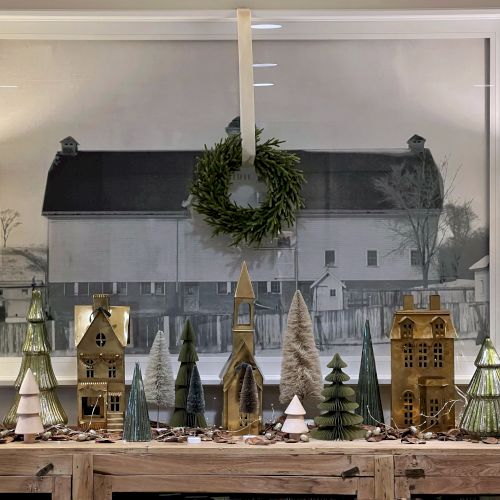 A winter holiday window display with a wreath, village houses, and evergreen trees under a large glass panel.
