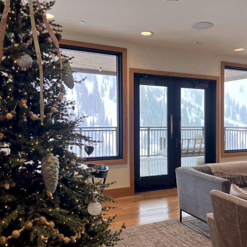 A decorated Christmas tree with warm lights fills an indoor lounge, with large windows showing snowy mountains and pine views outside.